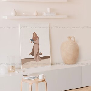 May include: A digital art print featuring a woman in a brown dress holding a blue bird. The print is framed and hanging on a white wall above a white shelf with books and decorative objects.