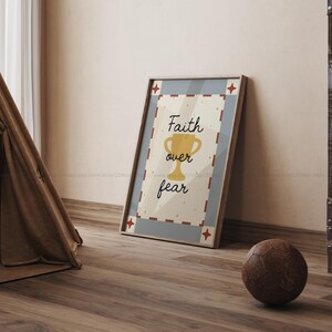 May include: A framed print with the words "Faith over fear" and a gold trophy illustration. The print has a light beige background with a blue border and red star accents. A brown leather soccer ball rests on the wooden floor.