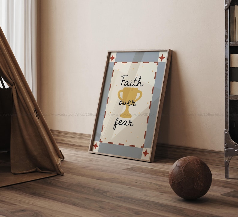 May include: A framed print with the words "Faith over fear" and a gold trophy illustration. The print has a light beige background with a blue border and red star accents. A brown leather soccer ball rests on the wooden floor.