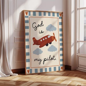 Könnte beinhalten: Gerahmter Kunstdruck mit einem roten Flugzeug, das zwischen blauen Wolken fliegt. Der Text "God is my pilot" ist in einer verspielten Schriftart geschrieben. Der goldfarbene Rahmen hat einen blau-cremefarbenen Rand.