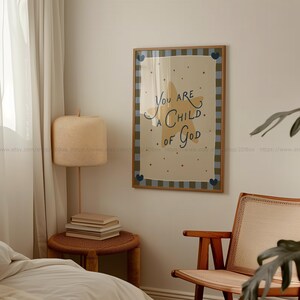 May include: A framed art print with the words "You are a Child of God" in blue script. The print features a star and heart design on a cream background, framed with a brown and blue checkered border. The print is displayed in a room with a lamp and chair.