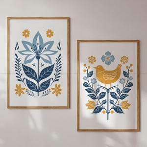 May include: Two framed art prints with botanical and bird designs. One print shows a large blue flower with leaves and yellow flowers. The other displays a yellow bird on a branch with blue and yellow floral accents. Both prints have a white background.