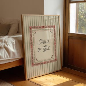 May include: A framed print with the words "Child of God" in elegant script. The print has a cream-colored background with a floral border and is set against a striped backdrop. The frame is a light wood color.