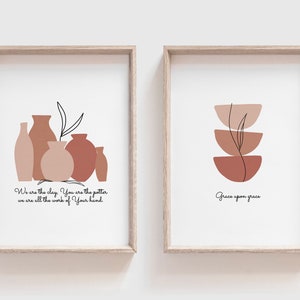 Puede incluir: Dos impresiones enmarcadas con diseños abstractos minimalistas en tonos marrones y rosados. La impresión de la izquierda presenta un grupo de jarrones con un solo tallo y una hoja. La impresión de la derecha presenta tres cuencos apilados con un solo tallo y una hoja. La impresión de la izquierda incluye el texto "We are the clay, You are the potter, we are all the work of Your hand." La impresión de la derecha incluye el texto "Grace upon grace."