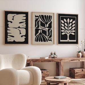 May include: Three framed abstract art prints. The prints feature black and cream designs: two doves, floral shapes, and a tree. The frames are a light wood colour. The art is displayed above a wooden console table and a white armchair.