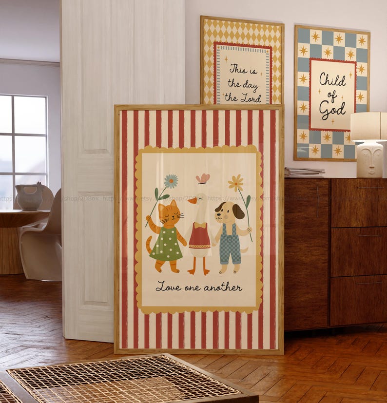 May include: A framed print with a red and white striped border features a cartoon cat, goose, and dog holding flowers. The text "Love one another" is below the characters. Two additional framed prints are visible, one with the text "This is the day the Lord" and the other with "Child of God".