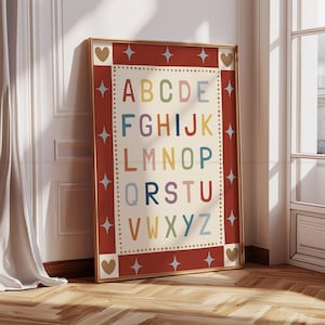 May include: A framed alphabet print with a red border and gold frame. The letters A through Z are displayed in various colors against a cream background. The border features star and heart accents. The print is leaning against a wall.