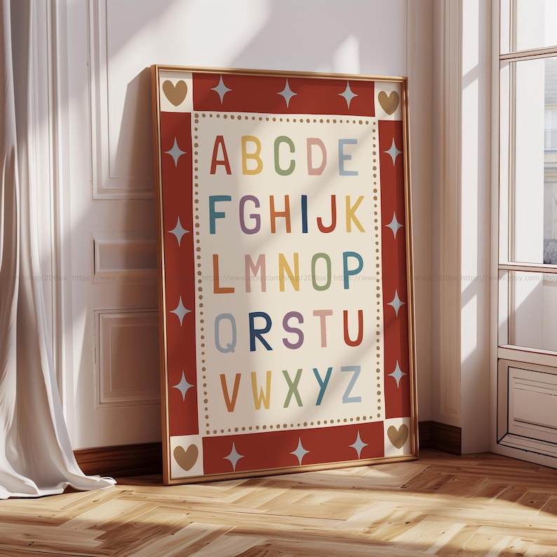 May include: A framed alphabet print with a red border and gold frame. The letters A through Z are displayed in various colors against a cream background. The border features star and heart accents. The print is leaning against a wall.