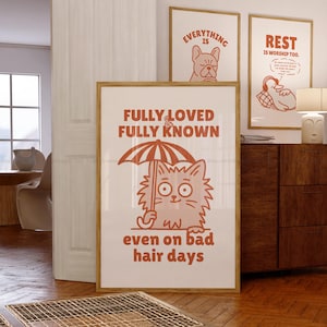 May include: Three framed art prints with a light wood finish. The largest print features a cartoon cat holding an umbrella with the text "FULLY LOVED & FULLY KNOWN even on bad hair days." The other prints have the text "EVERYTHING IS" and "REST IS WORSHIP TOO."