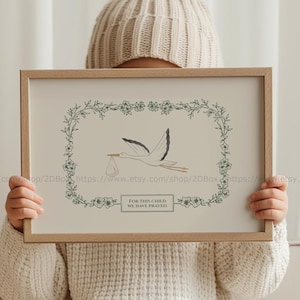Può includere: Una stampa incorniciata con un bordo floreale verde e una cicogna bianca che trasporta un bambino in una borsa. Il testo della stampa recita "Per questo bambino abbiamo pregato."