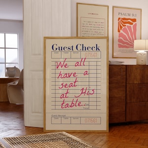 Op de afbeelding: Een bruin houten ingelijst bord met een witte achtergrond en zwarte lijnen. Het bord zegt "Guest Check" en heeft de tekst "We all have a seat at His table" geschreven in roze en blauw. Het bord heeft ook het nummer "071561" erop gedrukt.