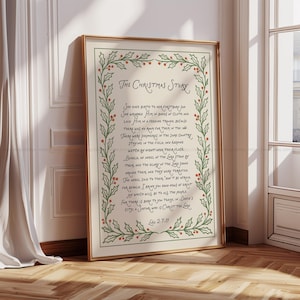 May include: A framed print with the text "The Christmas Story" in elegant script, surrounded by a green and red holly border. The text is centered on a cream-colored background, framed in gold.