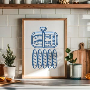 Könnte beinhalten: Ein gerahmter Druck mit einer blauen Illustration einer Sardinenbüchse, zwei Fischen und dem Wort "Sardines". Unter der Dose befinden sich fünf stilisierte Sardinen. Der Druck ist in einem Holzrahmen und in einer Küche ausgestellt.