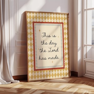 Puede incluir: Impresión enmarcada con el texto "This is the day the Lord has made". La obra de arte presenta un fondo crema con un patrón de diamantes y un borde rojo y crema. El marco es dorado.