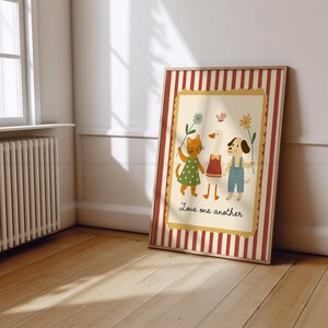 May include: A framed print featuring a cat, goose, and dog holding hands and flowers. The cat wears a green dress, the goose a red top, and the dog blue overalls. The print has a cream background with the words "Love one another". The frame has red and white stripes.