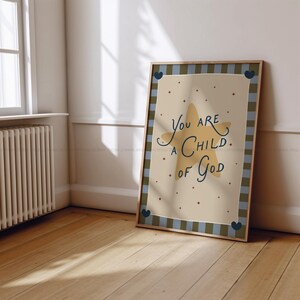 May include: A framed print with the text "You are a Child of God" in a whimsical font. The artwork features a large yellow star and small red dots on a beige background, framed by a blue and green plaid border. Decorative hearts are in each corner.