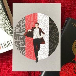 May include: A print featuring a figure in a long coat running in front of a circular design with red, gray, and black segments. The background is gray, and the print is on a white card. Books are visible on the sides.
