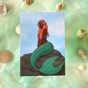 Könnte beinhalten: Postkarte mit einer Illustration einer Meerjungfrau. Die Meerjungfrau hat korallenfarbenes Haar, ein türkisfarbenes Oberteil und einen grünen Schwanz. Sie sitzt auf einem Felsen vor blauem Himmel. Muscheln und grüner Stoff umgeben die Postkarte.
