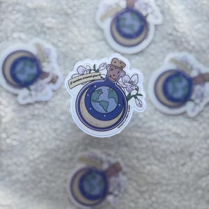 Könnte beinhalten: Eine Sammlung von Aufklebern mit einer blauen Zaubertrankflasche mit Korken, einem Mond und einer Weltkugel. Die Aufkleber haben ein Blumendesign und den Text "I revolve around you".