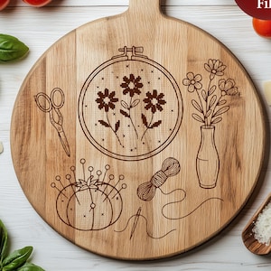 Op de afbeelding: Een ronde houten snijplank met een handvat, lasergegraveerd met een bloemenborduurontwerp, een schaar, een speldenkussen en een vaas met bloemen. De tekst "Laser Engraving File" staat op een rode cirkel.