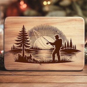 Pode incluir: Tábua de corte retangular de madeira clara com uma cena gravada de um pescador lançando sua linha ao pôr do sol. O design inclui um lago, árvores e um sol nascente, tudo em estilo rústico.