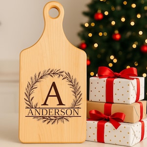 Könnte beinhalten: Ein helles Holzschneidebrett mit Griff und dekorativem Kranzdesign. Der Kranz umgibt den Buchstaben "A" und den Namen "Anderson". Das Brett wird neben verpackten Geschenken mit roten Bändern präsentiert.