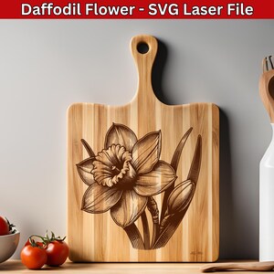 May include: A wooden cutting board with a laser-engraved daffodil flower design. The flower is brown and has a yellow center. The board is made of light wood and has a handle.