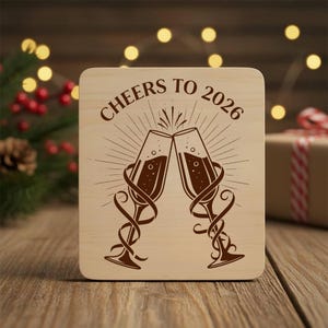 Könnte beinhalten: Ein quadratischer Untersetzer aus Holz mit dem Text "Cheers to 2026" und zwei Sektgläsern, die anstoßen. Das Design ist in Braun geätzt. Der Untersetzer befindet sich auf einer Holzoberfläche.