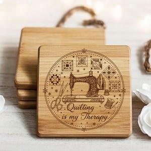 May include: Square wooden coasters with a laser-etched design. The design includes a vintage sewing machine, scissors, and the text "Quilting is my Therapy" within a circular frame. The coasters are made of light-coloured wood.