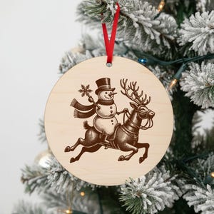 May include: A round wooden Christmas ornament with a snowman riding a reindeer. The snowman wears a top hat and scarf, holding a snowflake. The reindeer has a harness. A red ribbon is attached for hanging.