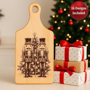 May include: A light-colored wooden cutting board with a handle, featuring a detailed engraving of two nutcracker figures and a Christmas tree. The board is accompanied by wrapped gifts with red ribbons. A red circle with the text "16 Designs Included" is also visible.