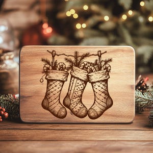 May include: A wooden cutting board with a laser-etched design of three Christmas stockings filled with candy canes and pine branches. The stockings are hanging from a string, and the board has a natural wood grain finish.