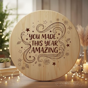 Könnte beinhalten: Runder Holzschild mit der Aufschrift "YOU MADE THIS YEAR AMAZING" in dekorativer Schrift. Das Schild hat Sterne und florale Akzente und ist aus hellem Holz gefertigt.