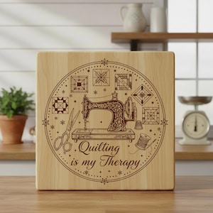 May include: A square wooden sign with a brown engraved design featuring a vintage sewing machine, scissors, and a spool of thread. The text "Quilting is my Therapy" is also engraved. The sign is light-coloured wood.