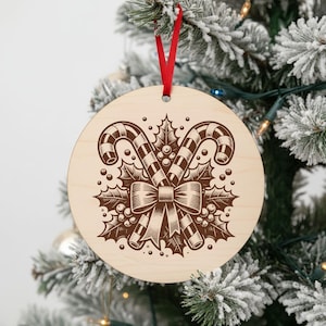 May include: Round wooden Christmas ornament with a red ribbon. The ornament features a brown engraved design of two candy canes, holly leaves, berries, and a bow. The ornament is hanging on a snowy Christmas tree.