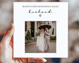 May include: A white card with the text "Happy First Birthday as my husband" and a heart symbol. The card features a photo of a couple walking down a street, holding an umbrella.