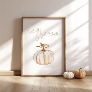 May include: A framed print featuring a watercolour pumpkin and the words "cosy season" in gold script. Two small pumpkins sit on the wooden floor in front of the frame. The large pumpkin in the print is white and tan.