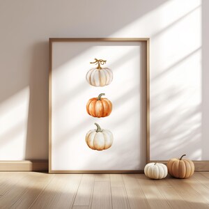 May include: A framed print showing three watercolour pumpkins in white and orange hues. The pumpkins are arranged vertically, within a wooden frame. Two small pumpkins are on the wooden floor in front of the frame.
