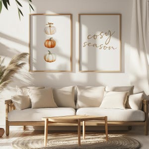 May include: Two framed art prints. One features three watercolor pumpkins in shades of white and orange. The other print has the words "cosy season" in gold script. The prints hang above a light-coloured sofa with cushions.