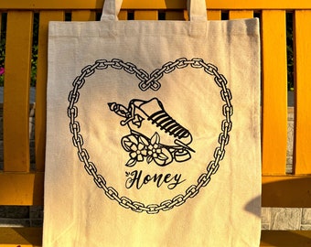 Könnte beinhalten: Eine weiße Canvas-Tragetasche mit schwarzem Druck. Der Druck zeigt eine Herzform aus einem Kettenglieddesign. Im Herzen befindet sich eine Schwarzweißillustration eines Rollschuhs mit Blumen und dem Wort "Honey".