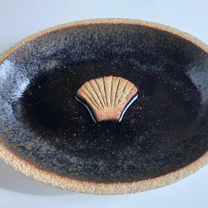 May include: An oval ceramic dish with a dark, speckled interior and a textured, light brown rim. A seashell-shaped accent with tan and black details is centered in the dish.