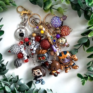 May include: A collection of keychains with various charms, including character figurines, beaded accents, and decorative baubles. The keychains feature a mix of colors, including red, silver, gold, and purple, with a variety of textures and shapes.