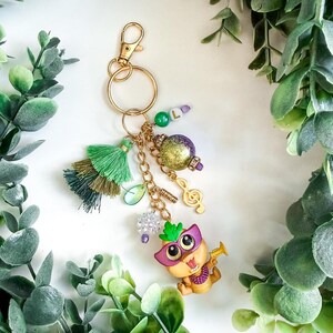 K&ouml;nnte beinhalten: Goldfarbener Schl&uuml;sselanh&auml;nger mit mehreren Anh&auml;ngern. Zu den Anh&auml;ngern geh&ouml;ren eine gr&uuml;ne Quaste, eine lila-goldene Perle, eine Musiknote und eine Zeichentrickfigur mit Maske und Trompete. Zus&auml;tzliche Perlen und ein Karabinerverschluss vervollst&auml;ndigen das Design.