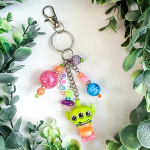 K&ouml;nnte beinhalten: Ein verspielter Schl&uuml;sselanh&auml;nger mit einer gr&uuml;nen Alien-Figur mit drei Augen, verziert mit bunten Perlen und Anh&auml;ngern. Der Schl&uuml;sselanh&auml;nger hat einen Karabinerverschluss und einen silbernen Schl&uuml;sselring. Die Perlen sind in Rosa-, Orange-, Blau- und Gr&uuml;nt&ouml;nen gehalten.