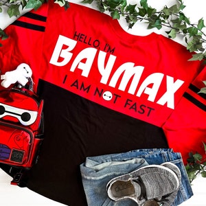 May include: Red and black jersey with the text "Hello, I'm Baymax I am not fast" in white. A white plush toy and a red backpack with a white face are in the image.