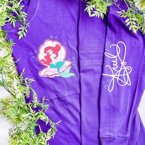 May include: Purple long-sleeved t-shirt with a graphic of Ariel from The Little Mermaid sitting in a seashell. The shirt has a white signature on the right sleeve.