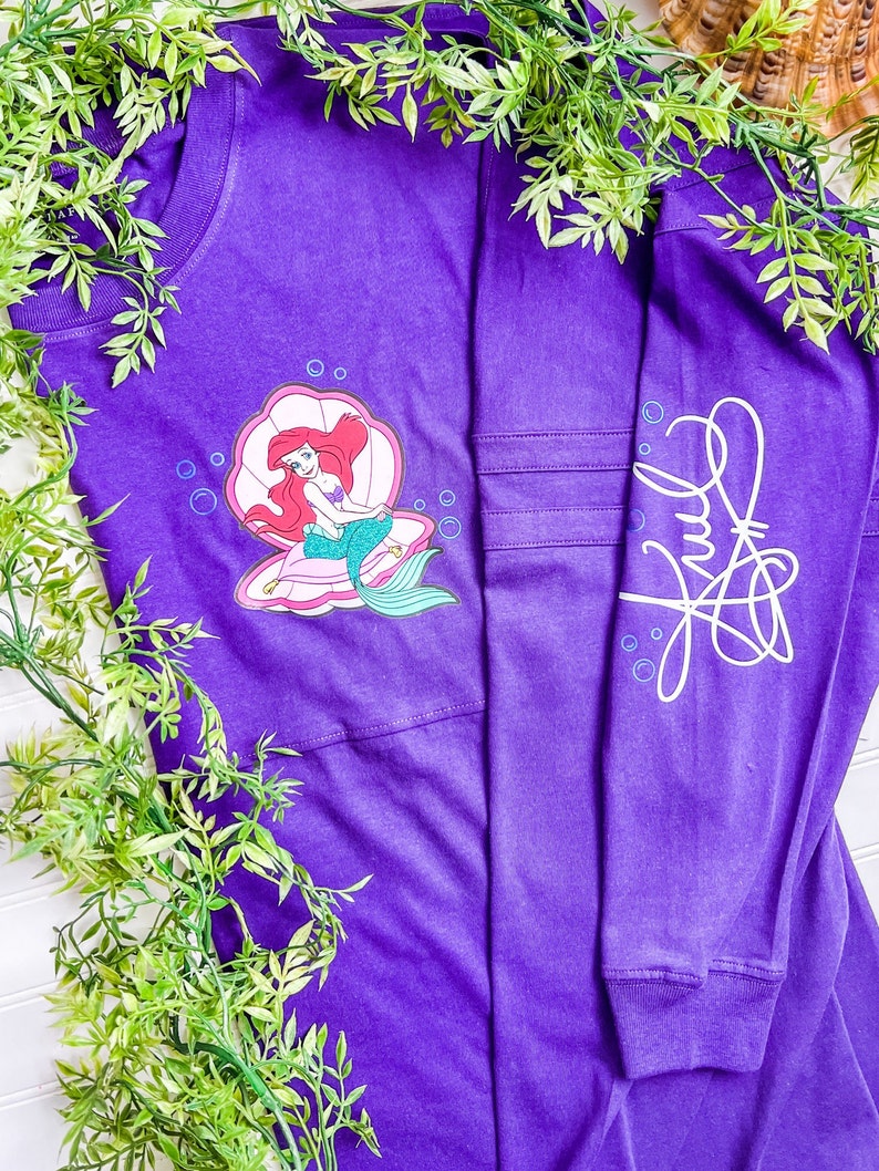 May include: Purple long-sleeved t-shirt with a graphic of Ariel from The Little Mermaid sitting in a seashell. The shirt has a white signature on the right sleeve.