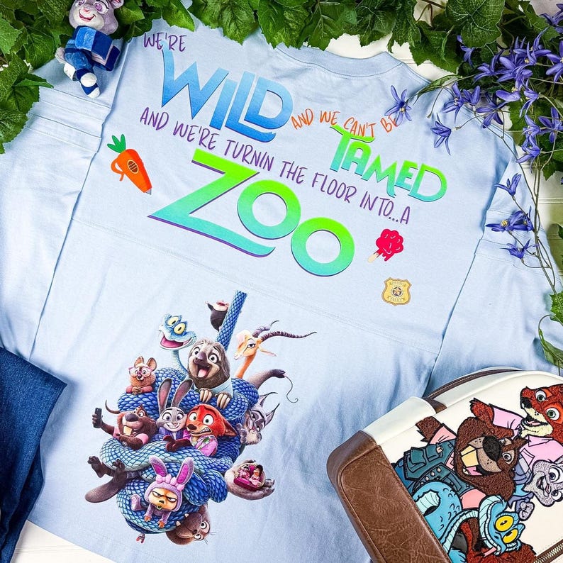 Puede incluir: Camiseta gr&aacute;fica azul claro con personajes de Zootopia. La camiseta tiene las palabras "We're Wild and We Can't Be Tamed" y "Zoo" en un dise&ntilde;o colorido. Tambi&eacute;n se ve una zanahoria y una insignia.