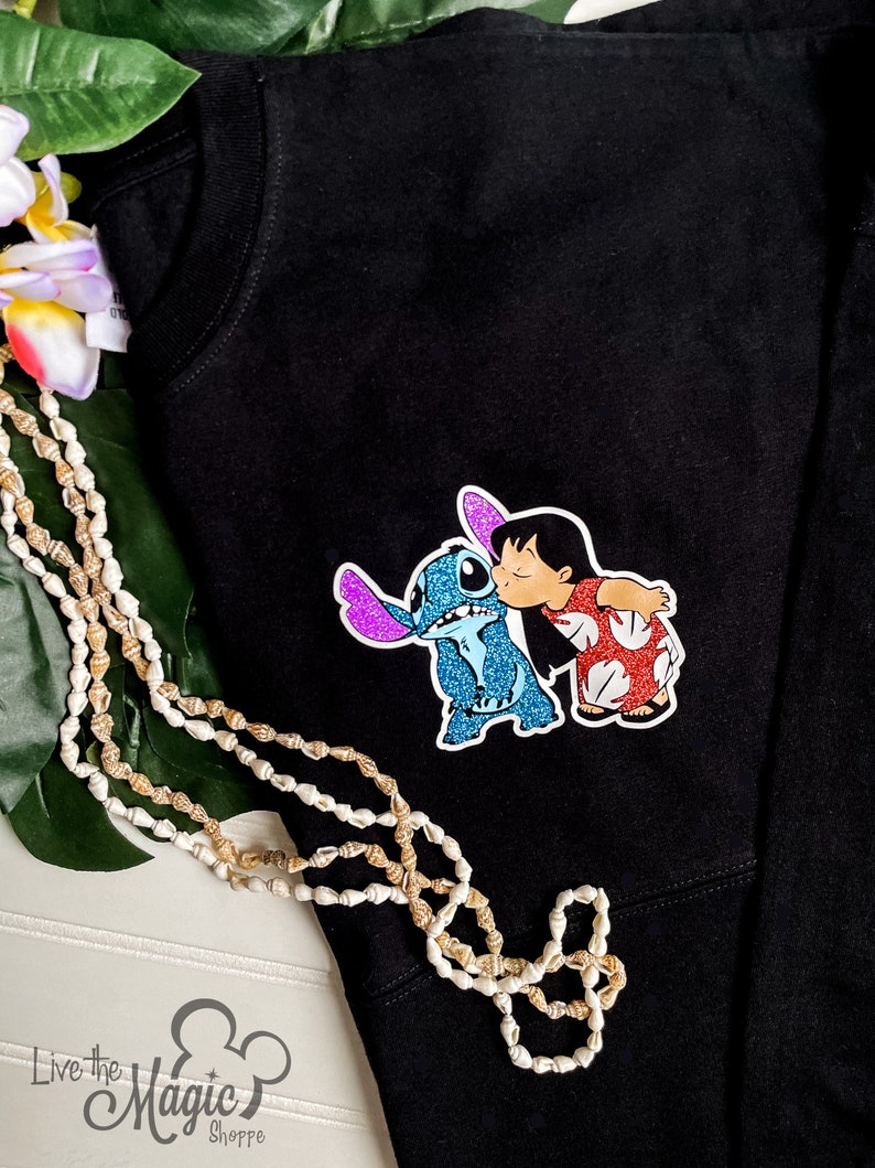 May include: Black t-shirt with a glittery blue Stitch and a red and white Lilo from the Disney movie Lilo & Stitch. The characters are kissing.
