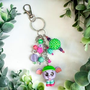 K&ouml;nnte beinhalten: Ein skurriles Schl&uuml;sselanh&auml;nger mit einer lila Elefantenfigur mit gr&uuml;nem Hut und bunten Perlenakzenten. Der Schl&uuml;sselanh&auml;nger enth&auml;lt einen silbernen Verschluss, eine Kette und verschiedene Perlen in Rosa, Gr&uuml;n und Lila.